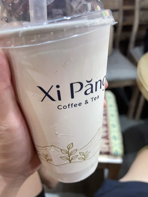 A close-up of a hand holding a clear plastic cup from 'Xi Pang Coffee & Tea' filled with a creamy white beverage.