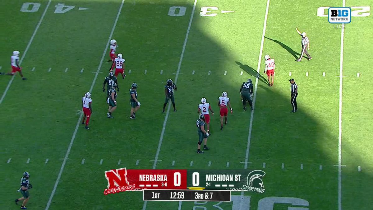 A wide view of a Big Ten Network broadcast shows Nebraska and Michigan State players regrouping between plays on the field, with the scoreboard displaying a 0-0 tie in the 1st quarter at 12:59 on a 3rd & 7. The wide shot captures the defensive and offensive players spaced across the green turf under bright sunlight, with referees nearby, fitting the post's note about a defensive play involving a blitz and an interception by Merritt.