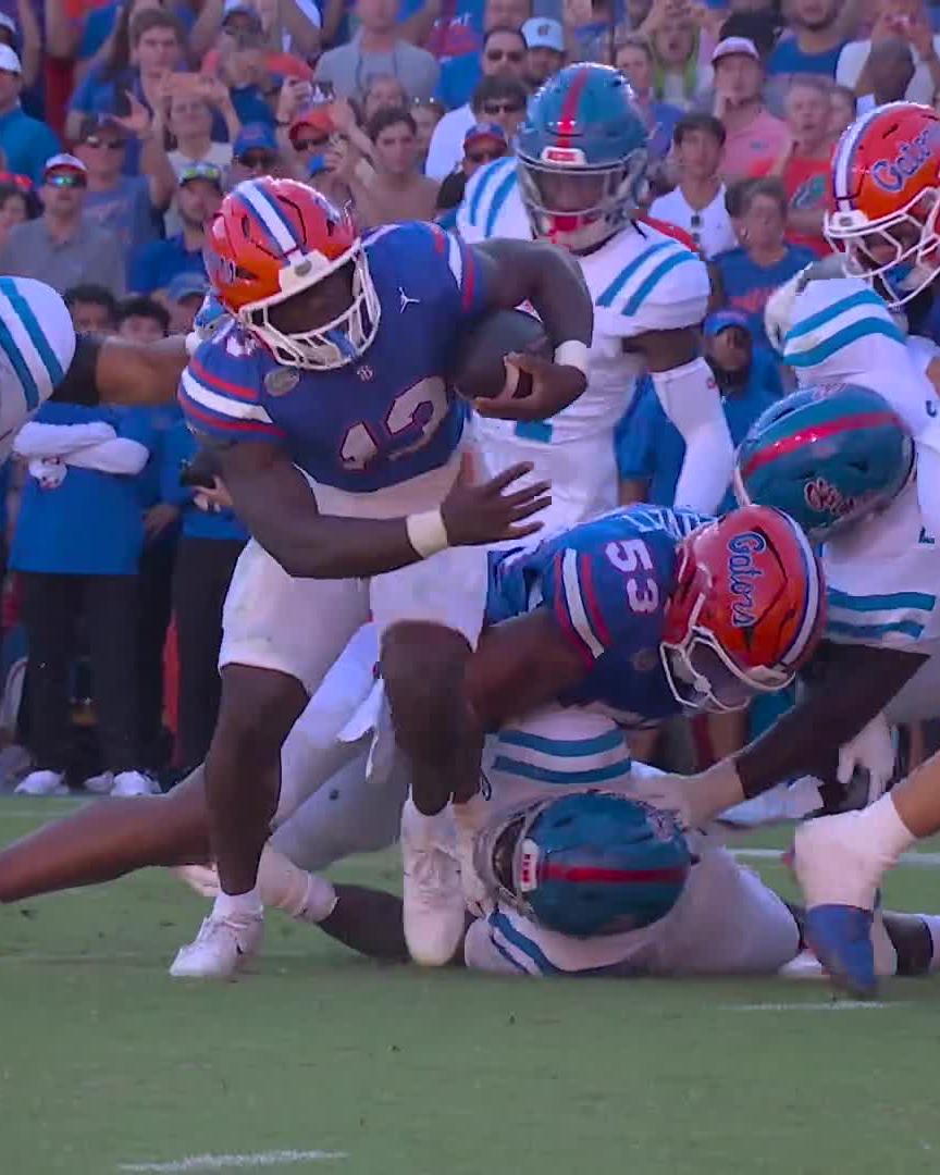 A Florida Gators player wearing #13 barrels through a group of Ole Miss defenders in blue and red helmets, charging down the field with the ball tucked away. The "CAN'T BRING BAUGH DOWN" caption perfectly matches the image, showcasing the powerful running back breaking tackles and refusing to be stopped by the opposing defense.