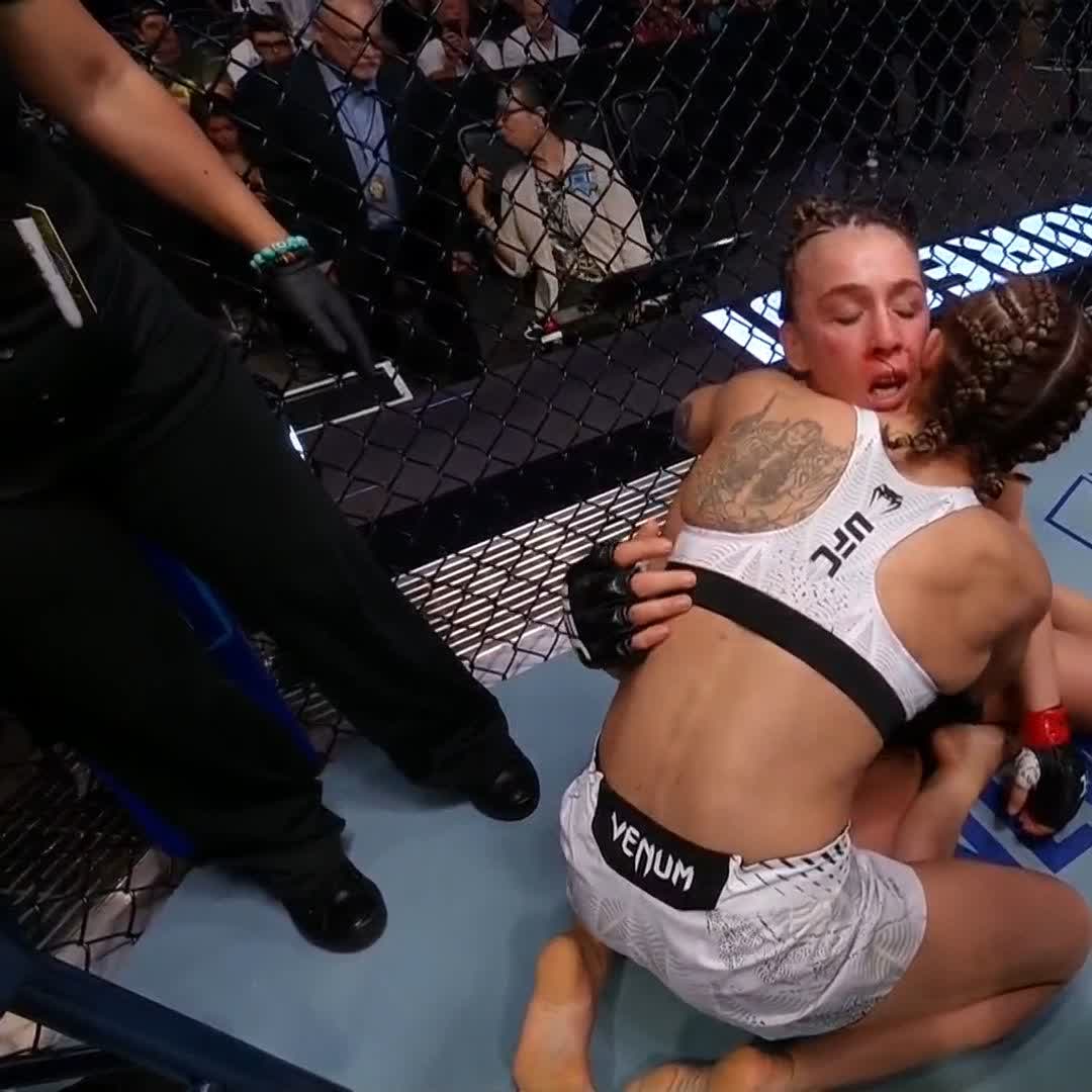 Two female UFC fighters in white Venum gear grapple on the mat near the cage during a match.