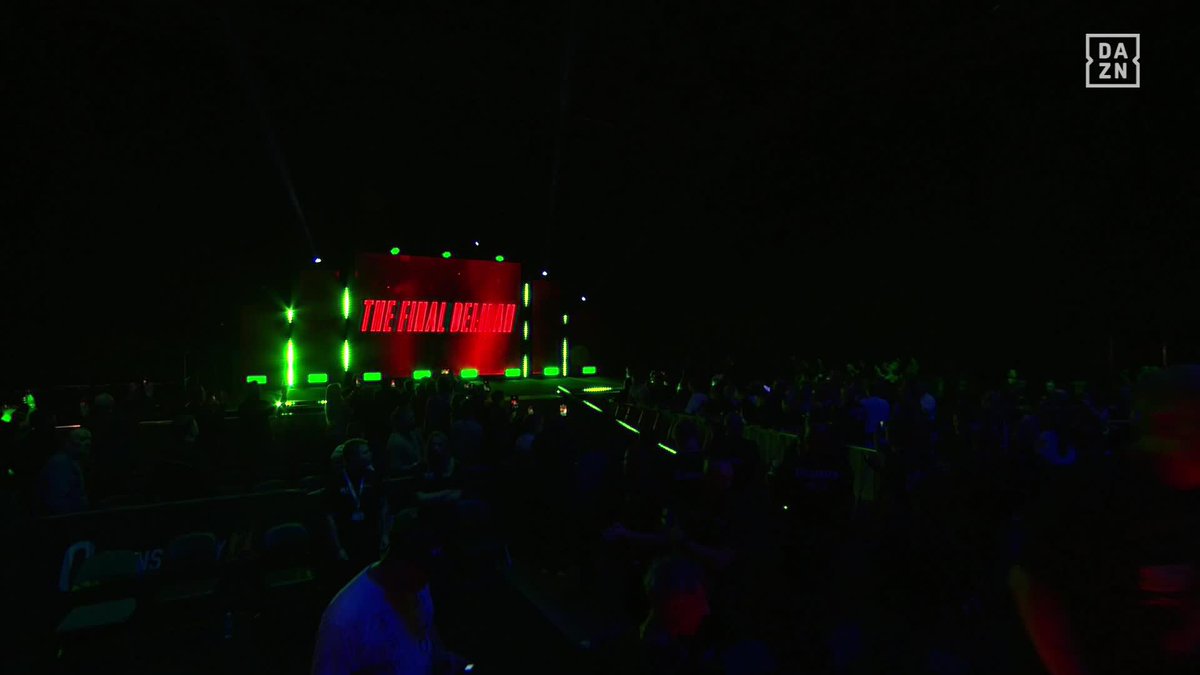 A dimly lit arena awaits the ring entrance, with a large screen illuminated in red and green lights displaying "THE FINAL HEADBAND," while spectators gather in the dark crowd below. The DAZN logo appears in the top right corner, tying into the boxing event promotion for the Heaney vs. Warburton fight on the streaming platform.