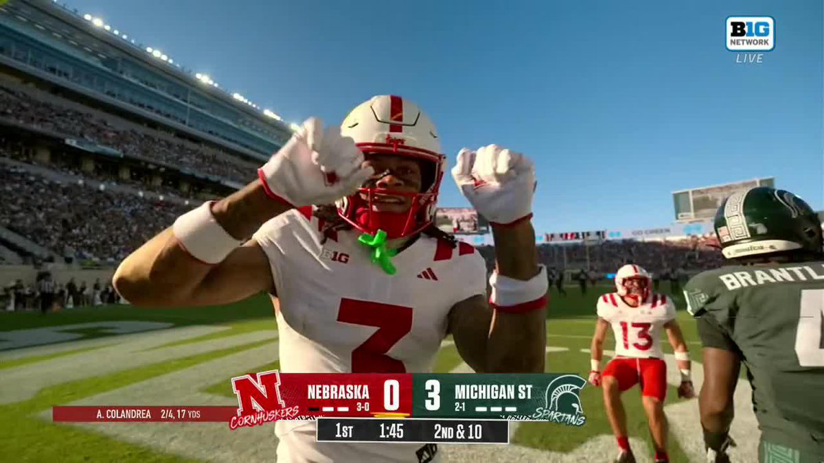 Nebraska wide receiver Kwazi Gilmer (#7) flexes toward the camera after a remarkable catch, wearing his white Cornhuskers jersey and helmet, with the scoreboard showing Michigan State leading Nebraska 3-0 in the 1st quarter with 1:45 remaining on a 2nd & 10 play, while teammate #13 and a Spartan defender (#5 Brantley) stand in the background at Spartan Stadium.