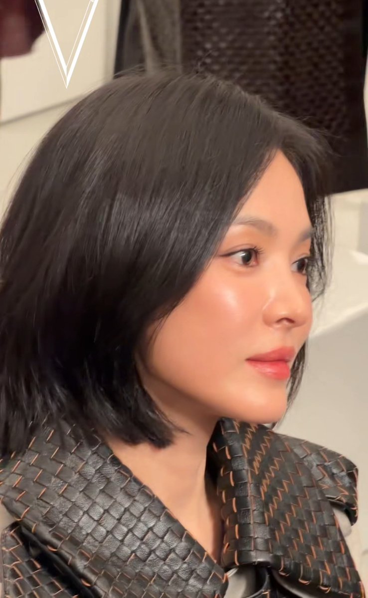 A close-up selfie of a woman with shoulder-length dark hair wearing a black woven leather top with orange stitching.