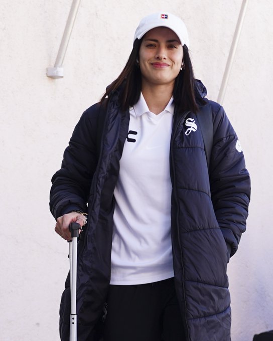A smiling woman wearing a white cap with "TYR" branding, a white polo shirt featuring the "SS" logo, and a dark puffer Moncler jacket pulls a rolling suitcase, arriving at Estadio Hidalgo as part of the Tuzas squad for the Liga Femenil BBVA match against Pachuca.