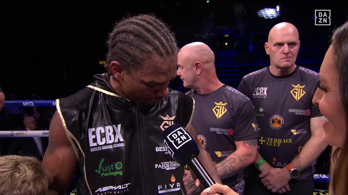 A boxer wearing a black "ECBX" vest and cornrow braids speaks into a DAZN microphone while looking down emotionally, surrounded by his team in a boxing ring.