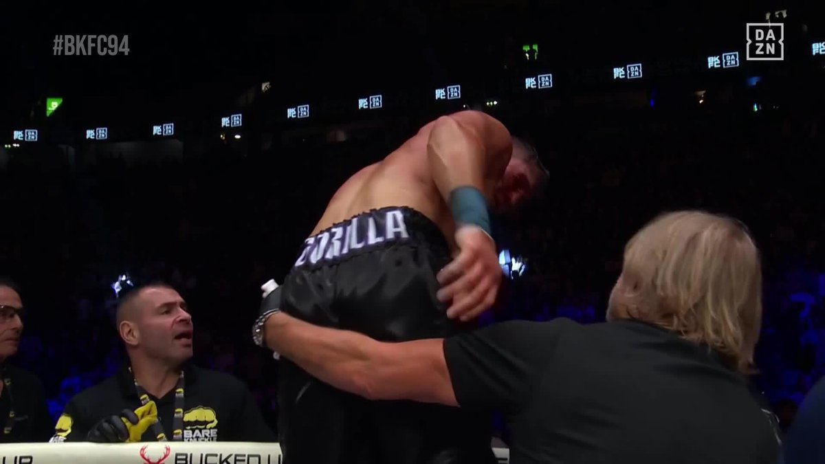 A shirtless fighter wearing black "GORILLA" shorts stands in the ring at #BKFC94 as a referee in black grabs his arm, apparently after his unanimous decision victory. The DAZN and PIES 96 branding is visible around the arena, with a cornerman wearing yellow gloves looking on from the side.