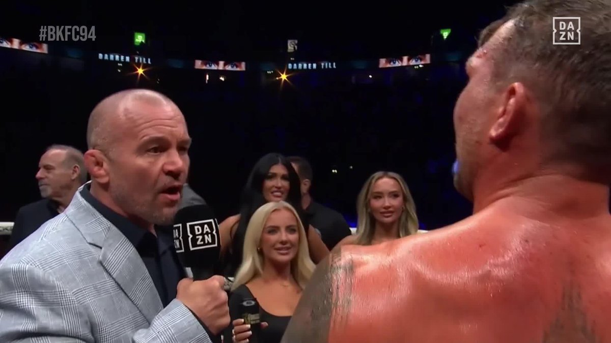 A bearded interviewer in a gray plaid blazer holds a DAZN microphone while speaking to a sweaty, shirtless fighter, with "BKFC94" and "DARREN TILL" displayed on arena screens behind them. Several women stand between the two men, and a "DAZN" logo appears in the upper right corner, capturing the post-fight confrontation moment referenced in the caption.