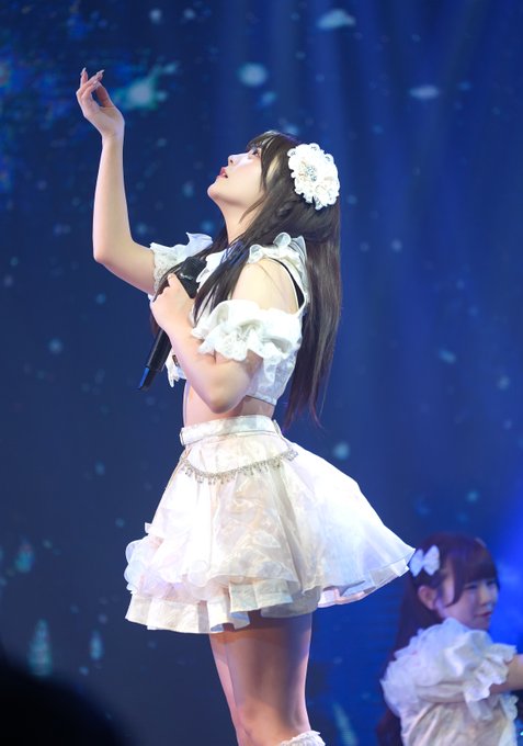 A concert photograph of a young woman in a white ruffled idol outfit with a white flower hair accessory, holding a microphone and reaching upward on a blue-lit stage.