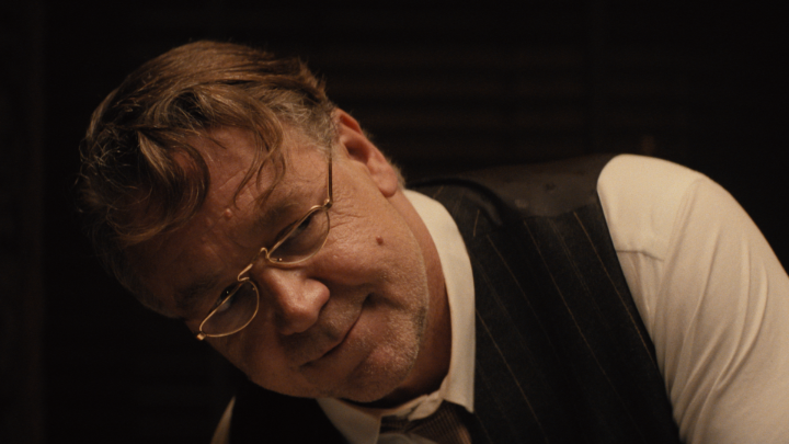 A man with glasses, wearing a white shirt and dark pinstriped vest, looks downward with a serious expression in a dimly lit setting.