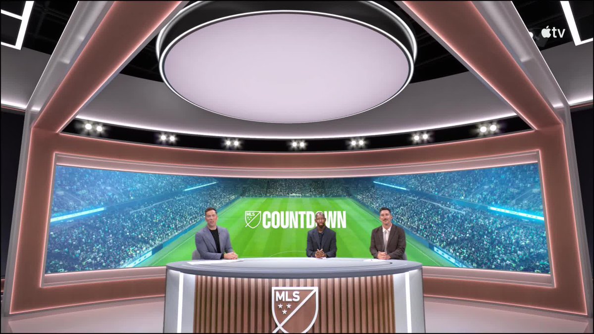 A studio shot of the MLS Countdown broadcast on Apple TV, featuring three presenters seated at a desk in front of a large screen displaying a soccer stadium.