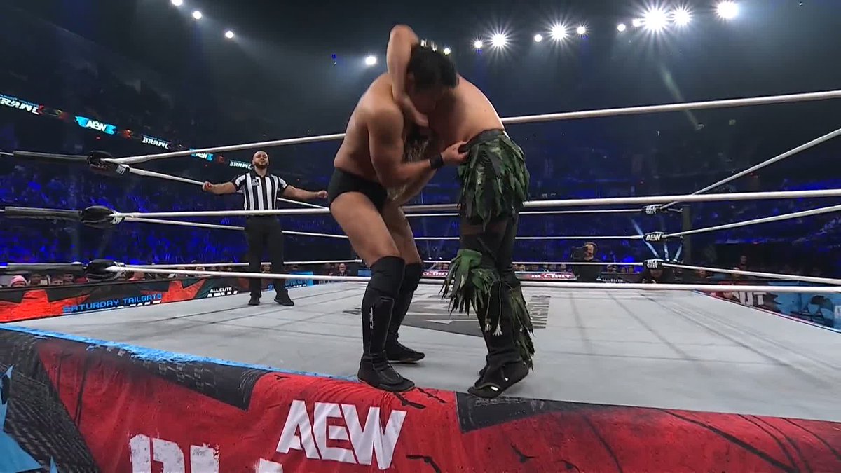 Two AEW wrestlers grapple inside the ring while a referee observes, with "AEW" branding visible on the apron and "Saturday Tailgate Brawl" signage displayed around the arena. The image captures an in-ring struggle during the AEW Saturday Tailgate Brawl event, showing one wrestler being planted into the apron area as referenced in the post's commentary about the match.
