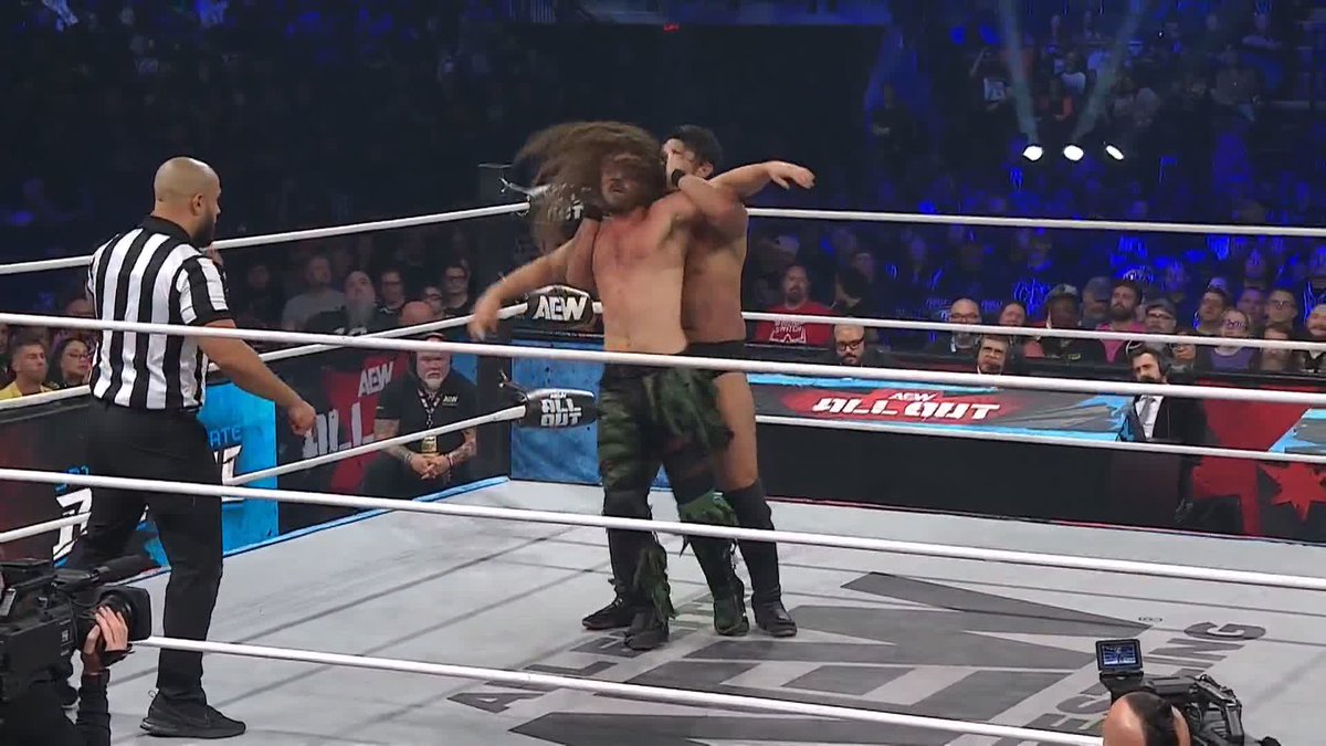 Two professional wrestlers engage in a grappling move inside a wrestling ring during an AEW event, with a referee in black and white stripes observing and a crowd watching from the arena seats.