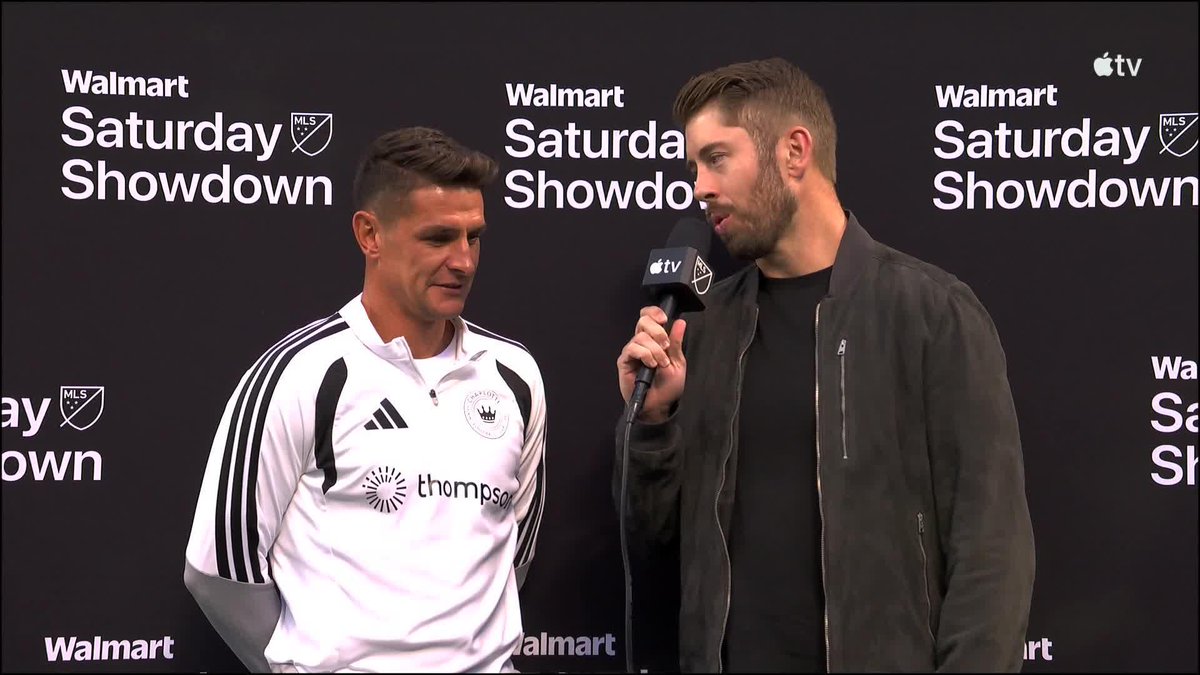 A reporter holding an Apple TV microphone interviews a Charlotte FC player in a white Adidas jersey with a Thompson sponsorship logo, with the backdrop displaying "Walmart Saturday Showdown" and MLS branding, relating to Ashley Westwood's post-game comments about teammate Allan Saint-Maximin.