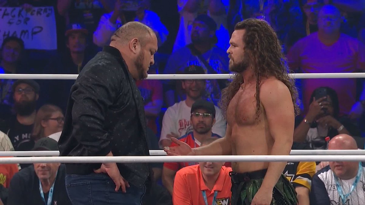 Two professional wrestlers face off inside a wrestling ring, with a bearded man in a black jacket and jeans on the left extending a hand toward a shirtless, long-haired wrestler in green pants on the right, amid an energetic crowd holding signs. The post about Samoa Joe delivering a headbutt to Jack Perry aligns with this tense post-match or pre-match confrontation between the two competitors.