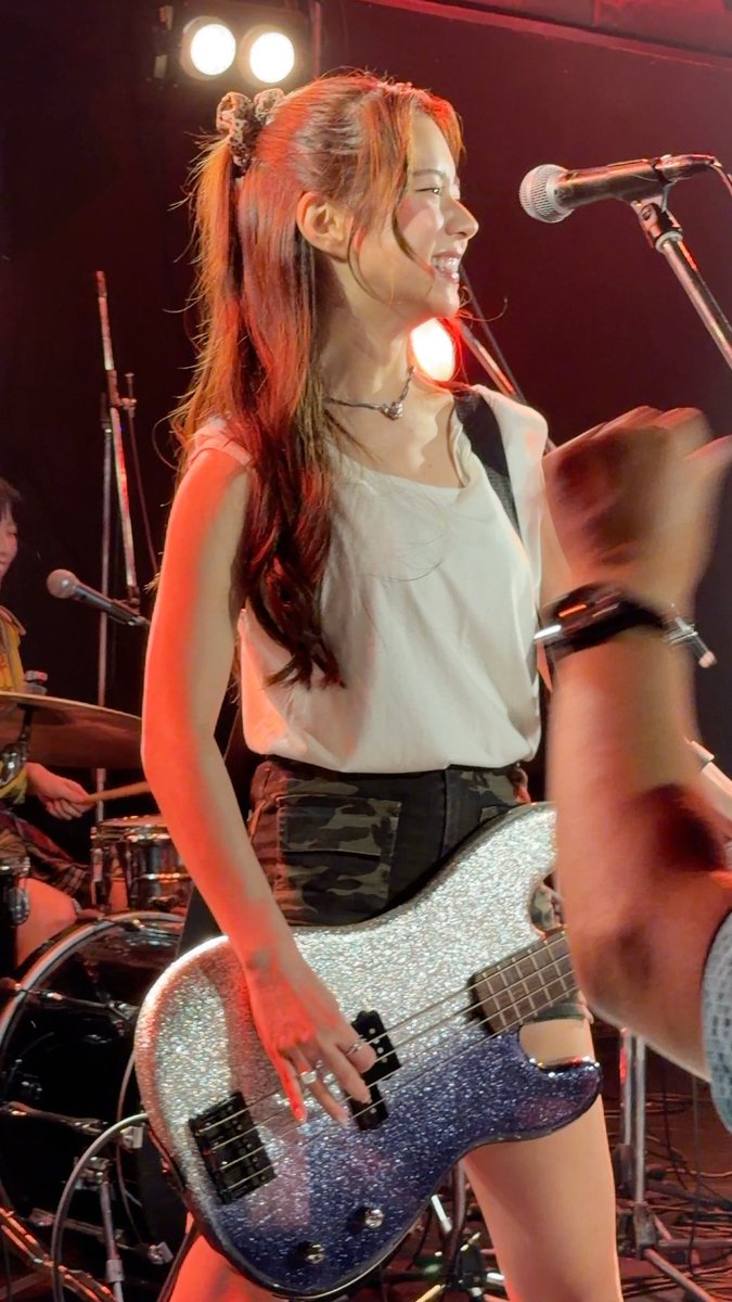 A young woman with long reddish-brown hair tied in a high ponytail performs live on stage, playing a sparkling silver glitter bass guitar while smiling into the microphone, wearing a white tank top and camouflage shorts. The energetic live concert scene perfectly captures the band's live performance at the Music City Tenjin festival in front of Fukuoka City Hall.