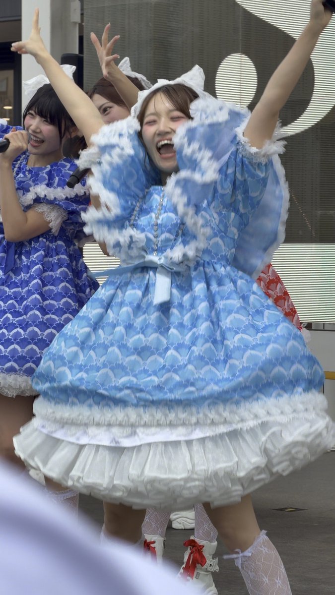 A group of Japanese idol performers in blue and white frilly dresses energetically performing, with one in the foreground smiling widely with her arms raised.