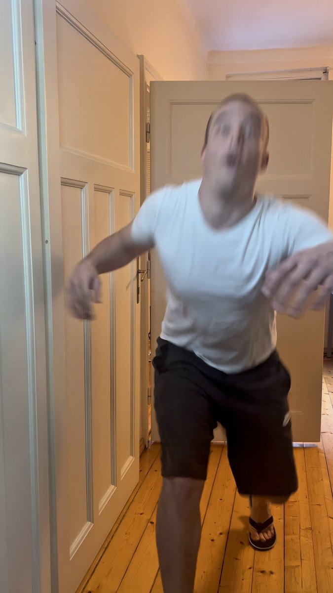 A blurry, low-angle shot shows a man in a white t-shirt and black shorts lunging toward the camera in a hallway with wooden floors and white doors.