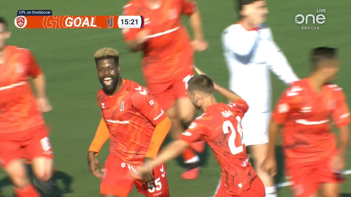 A Forge FC player celebrates with teammates after scoring the opening goal on a set-piece, as shown by the "GOAL" graphic and 15:21 match clock displayed on the OneSoccer broadcast. The jubilant player with bleached blond hair and beard smiles broadly while wearing the team's red jersey featuring sponsor logos and the number 55.