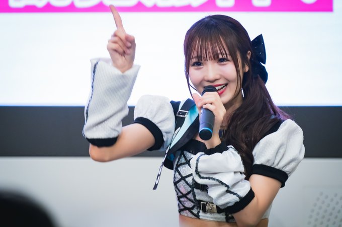 A smiling woman in a black and white maid-themed outfit holds a microphone and points upward on stage.