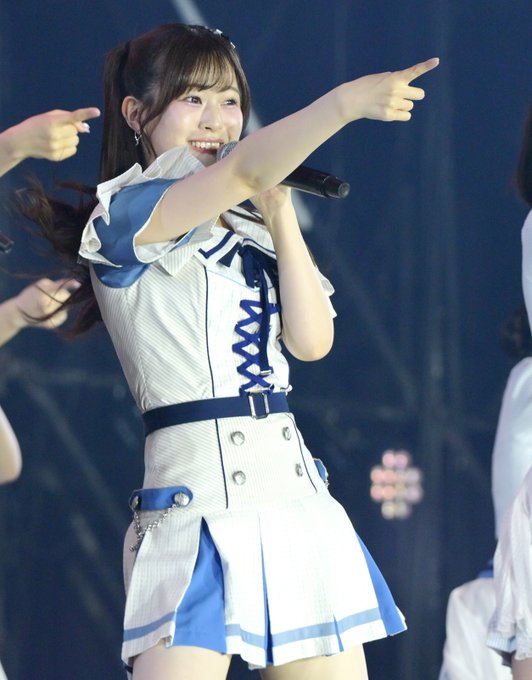 A young woman in a white and blue stage outfit with a bow points outward while singing into a microphone during a live performance.
