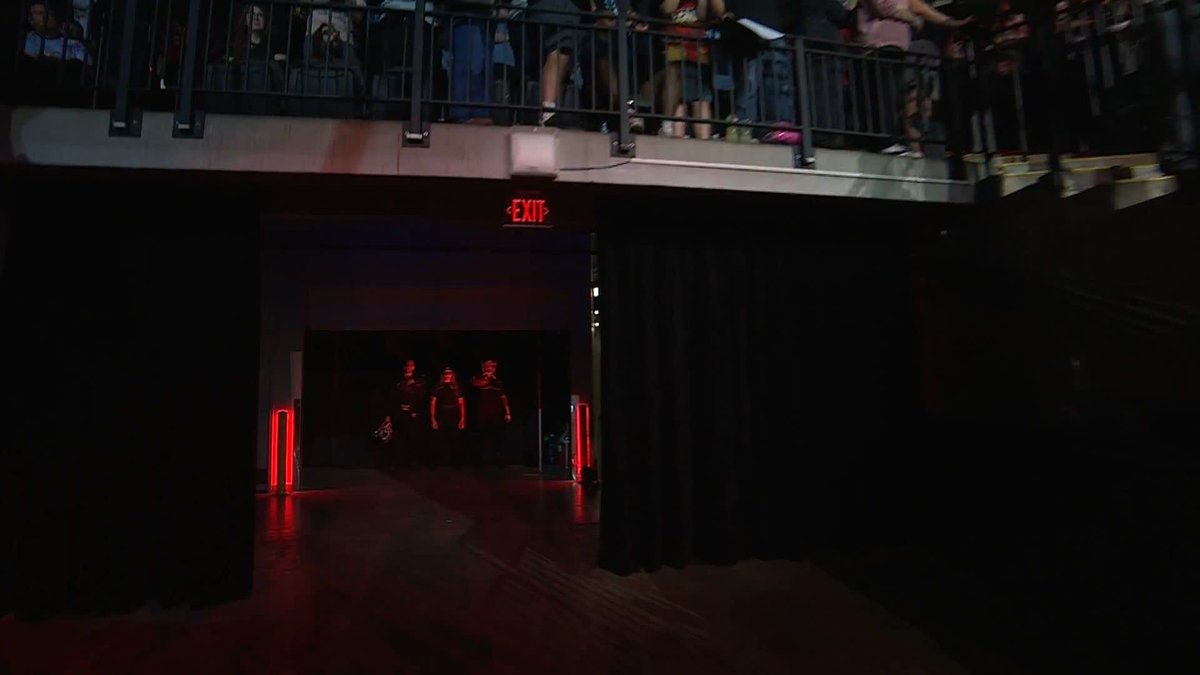 Three wrestlers make their dramatic entrance through a dark tunnel illuminated by red lights and an "EXIT" sign overhead, while fans watch from a balcony above. The image perfectly captures the pre-match atmosphere as the Death Riders prepare to engage in the AEW Tailgate Brawl event.