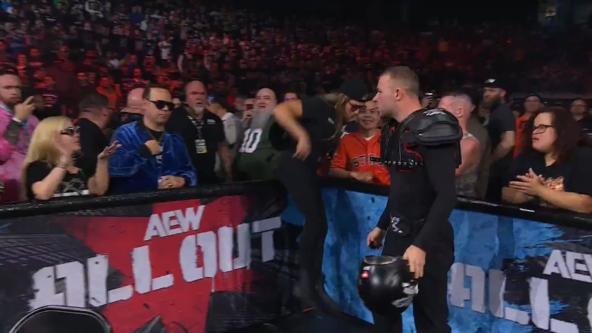 Daniel Garcia, wearing black shoulder pads, holds a helmet and shouts back at a fan from inside the ring during the AEW Tailgate Brawl, as Marina Shafir (visible in the crowd) escalates their rivalry by shouting at him from ringside.