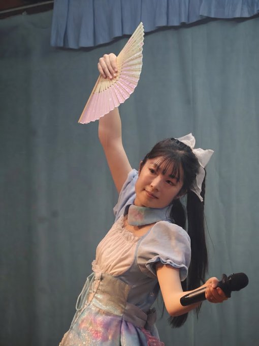 A performer in a pastel blue and pink outfit holds up a folding fan while performing with a microphone on stage.