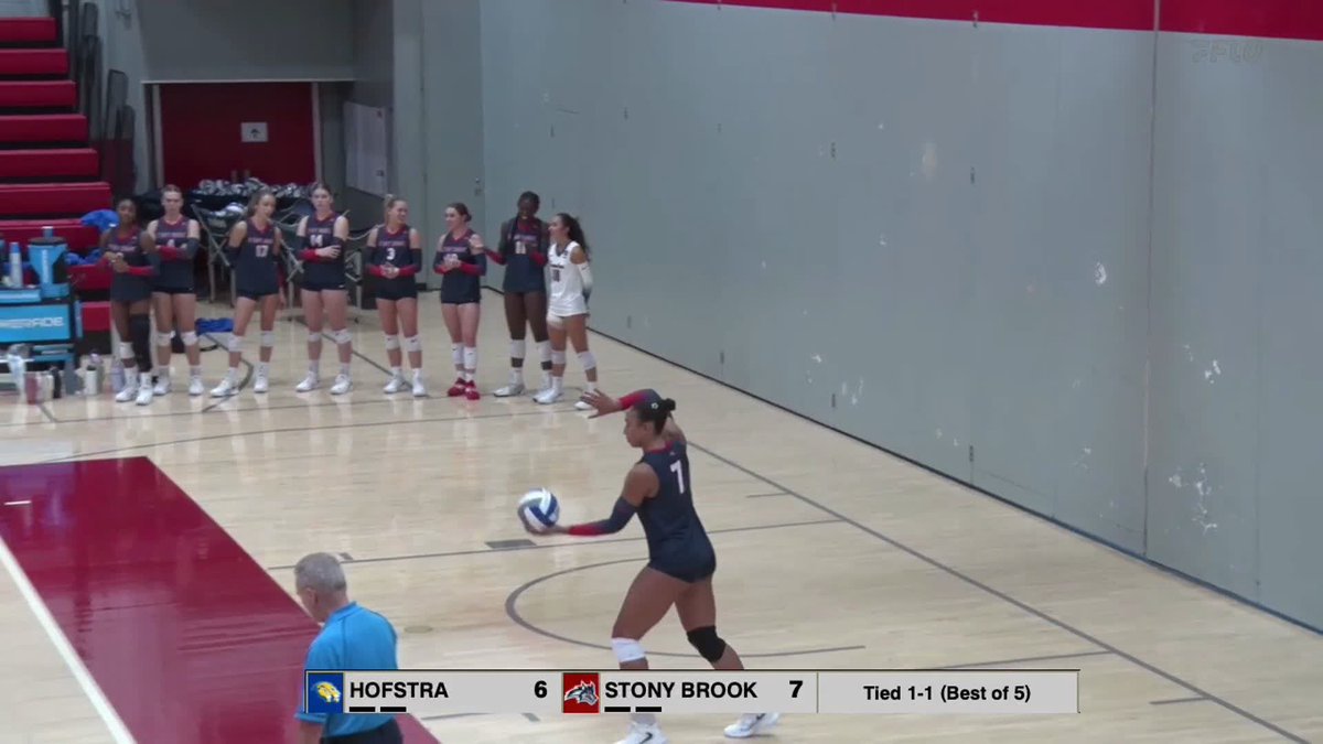 A volleyball player wearing number 7 prepares to serve the ball while her teammates in matching uniforms stand in a line along the court. The scoreboard at the bottom reads 'HOFSTRA 6, STONY BROOK 7, Tied 1-1 (Best of 5).'