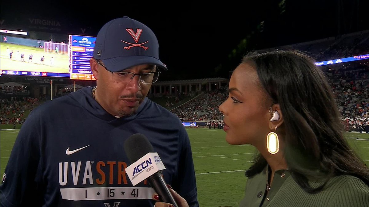 A post-game sideline interview at a University of Virginia football game, showing a coach wearing a UVA hat and jacket being interviewed by a reporter holding an ACCN ESPN microphone.