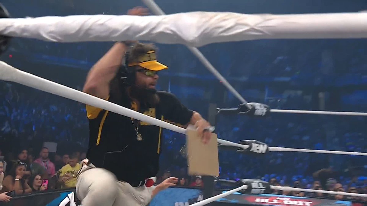 A wrestler with long hair, sunglasses, and a yellow visor leaps through the ropes of an AEW ring while holding a box, as the "ALL OUT" logo is visible on the ring apron—capturing the chaotic moment that led to the show being shut down.