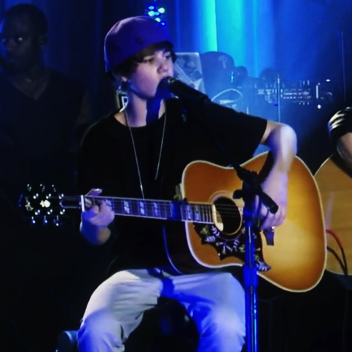 A young musician wearing a blue beanie and black t-shirt plays an acoustic guitar and sings into a microphone on a stage lit with blue lights.