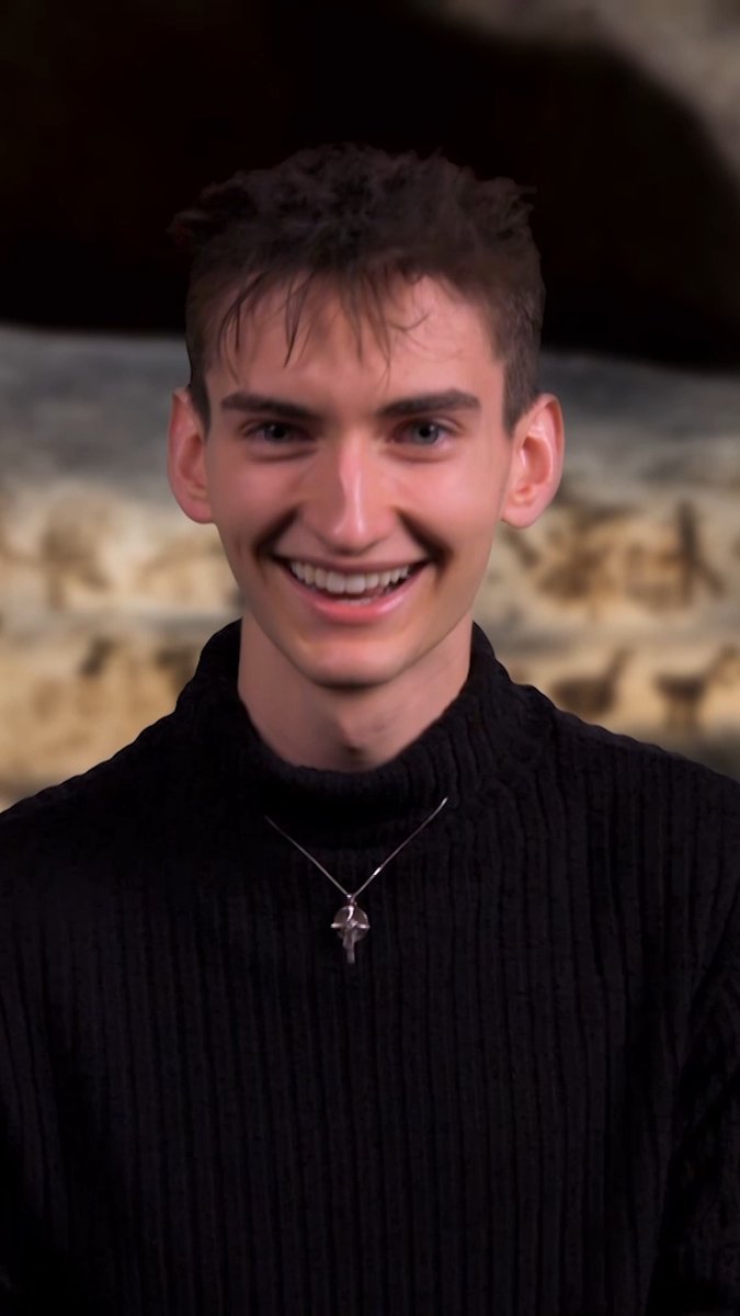 A young man with short dark hair smiles brightly while wearing a black turtleneck sweater and a silver cross necklace.