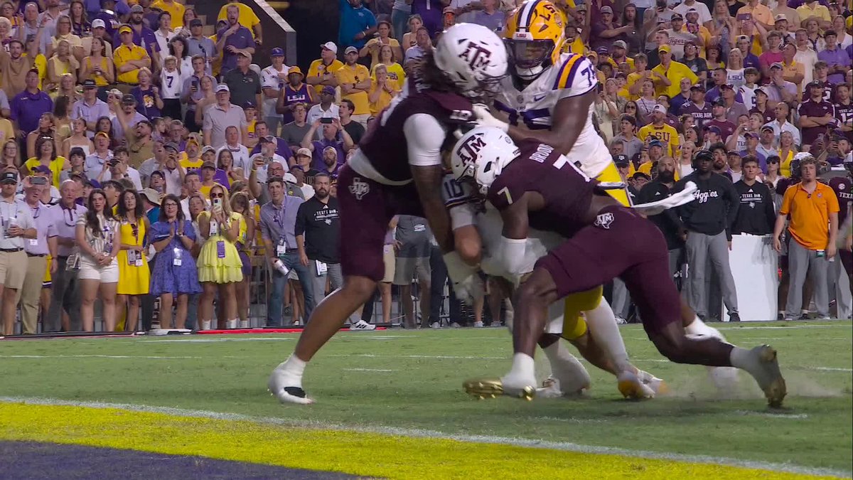 Two Texas A&M defenders in maroon jerseys, including #7, tackle an LSU player in a white and gold jersey (#71) on the field after what appears to be a touchdown, with the crowd watching from the stands behind them. The image relates to the post as it captures the immediate aftermath of LSU scoring first, with the energetic crowd and sideline personnel reacting to the play.