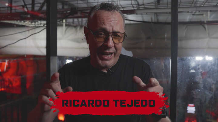 A man with glasses and short hair speaks animatedly with his hands raised, with a bold red banner displaying the name "RICARDO TEJEDO" overlaid across the bottom. The dramatic, dimly lit industrial-style background with red lighting gives the scene a theatrical feel, inviting viewers to guess who he resembles based on the prompt.