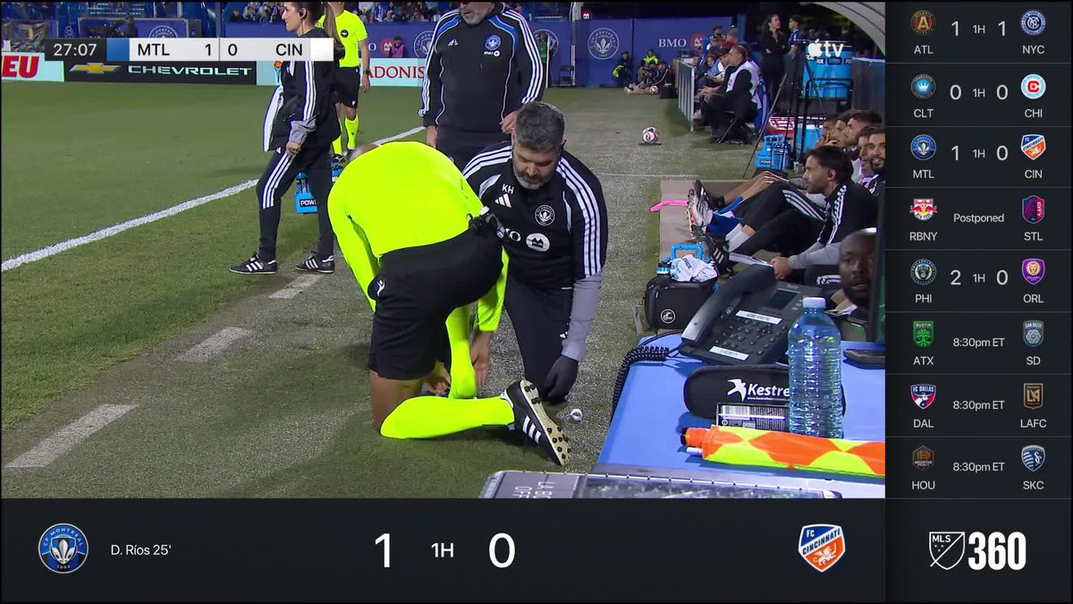 A soccer player in a bright yellow uniform crouches on the sideline while a medical staff member in a black tracksuit tends to him, with the scoreboard showing MTL leading CIN 1-0 at 27:07. Other MLS fixtures are listed on the right sidebar, including ATL vs NYC and PHI vs ORL 2-0, suggesting the referee required medical attention during the match.