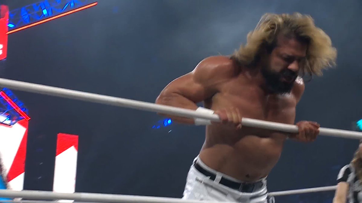 A shirtless, muscular wrestler with long blonde hair and a beard grips the top ring rope, appearing fatigued during an AEW match. The scene aligns with the post referencing the National Champion facing off against @BastardPAC and @MarinaShafir at #AEWAllOut.