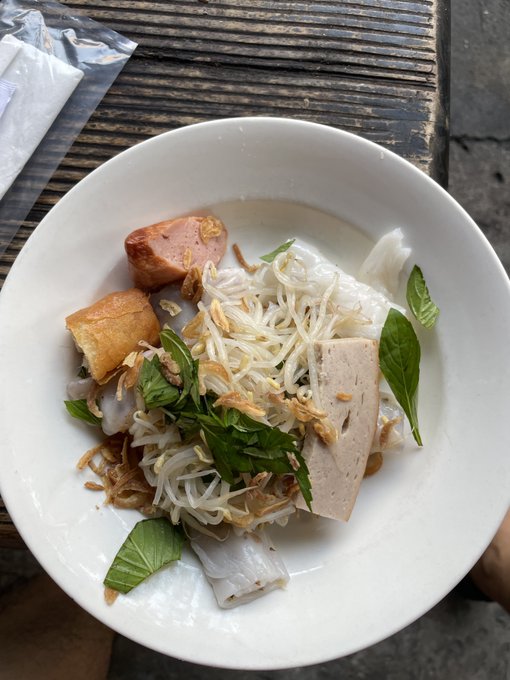 A white bowl sits on a slatted wooden table containing a noodle dish garnished with fresh green leaves, crispy fried shallots, a slice of sausage, fried dough, and rectangular pieces of meat or tofu.