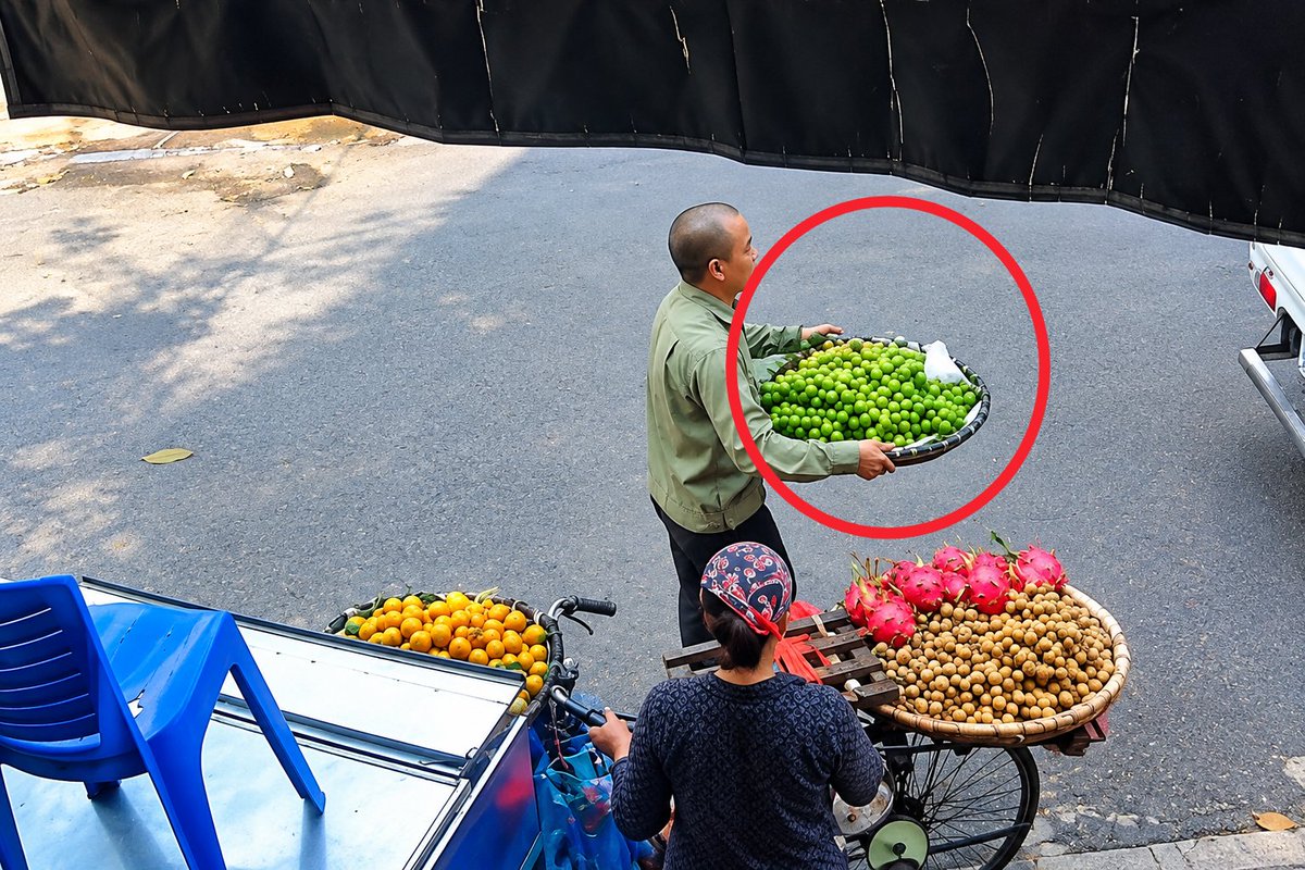 A man in a green shirt carries a large round tray overflowing with small green citrus fruits, likely limes, past a street vendor woman selling dragon fruit, longan, and oranges from her bicycle cart on a roadside. The scene captures a moment of street commerce, where the man's tray of fresh produce is highlighted in red, suggesting it plays a central role in the unfolding story of the roadside vendor confronting authorities.