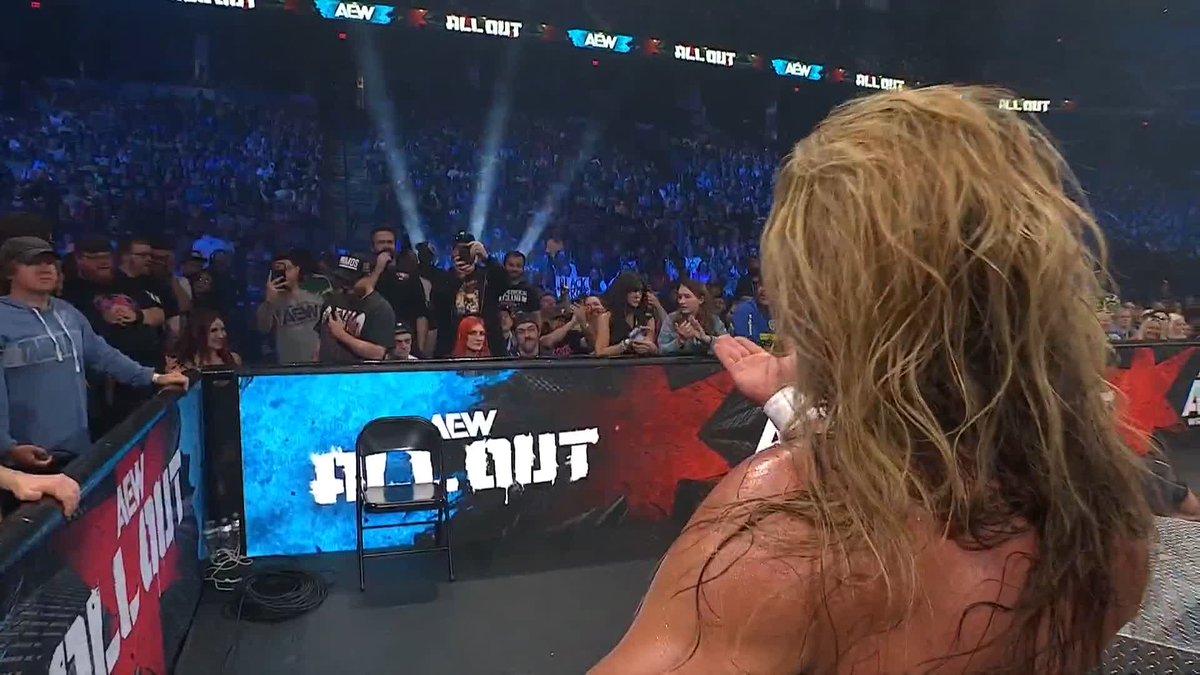 A view from behind a sweaty wrestler with long blonde hair standing in the ring at an AEW 'ALL OUT' event, looking out at a cheering crowd.