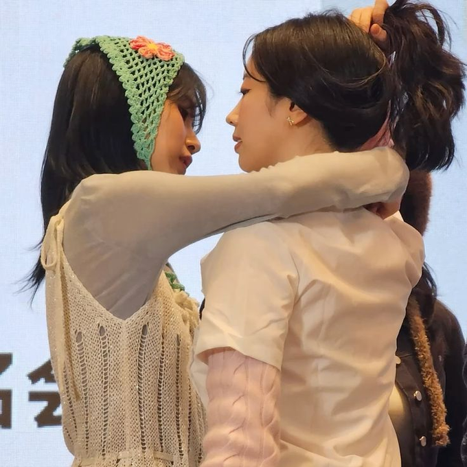 Two young women embracing closely face-to-face at what appears to be an event, one wearing a green crocheted headband with flowers and a cream knit top, the other in a white blouse with her hair in a ponytail.