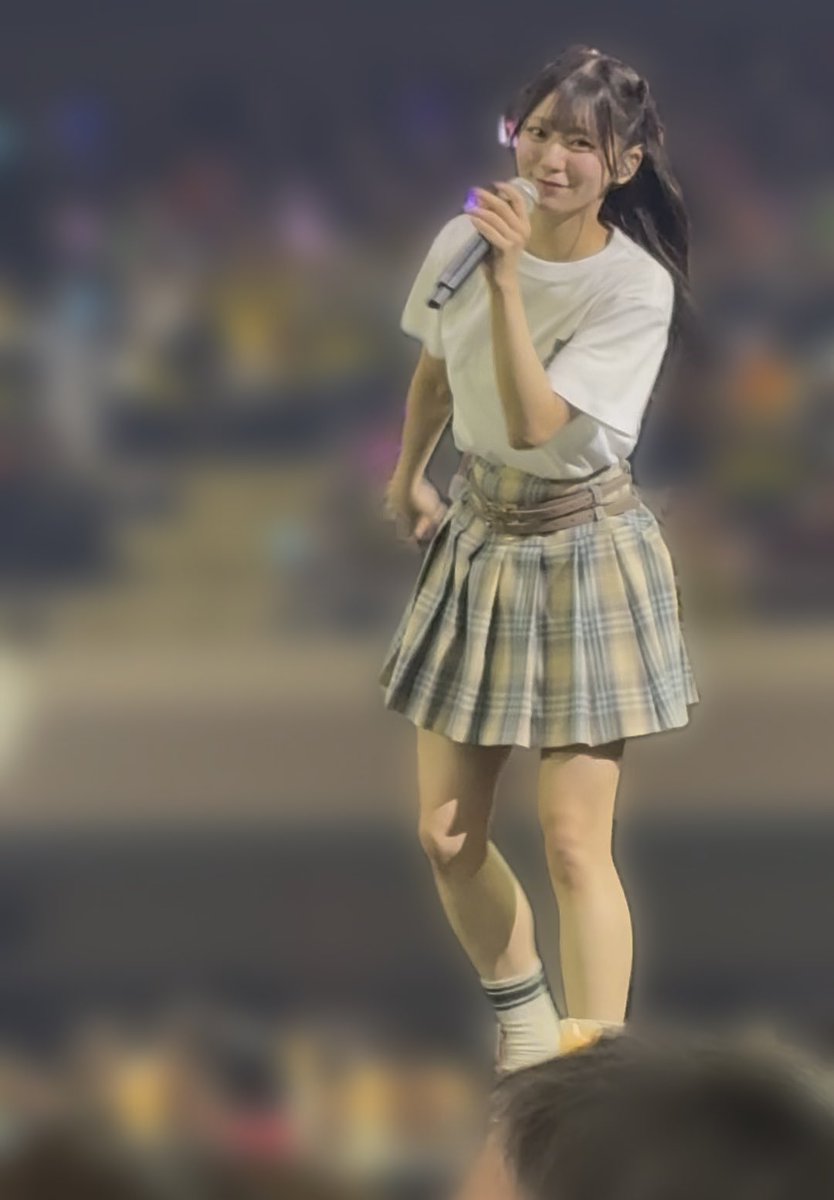 A young woman stands on stage holding a microphone, wearing a white t-shirt and a beige and green plaid pleated skirt with a brown belt, smiling at the audience.