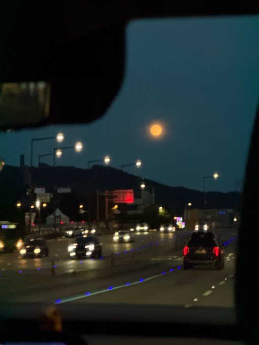 A view through a car windshield at dusk shows a highway with traffic, streetlights, and an orange moon rising over distant hills.
