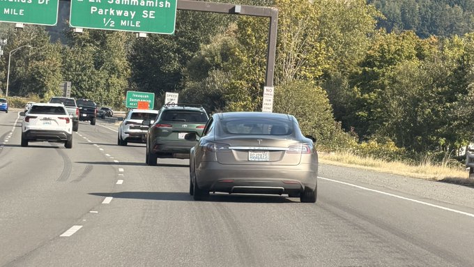 A highway scene with a gray Tesla sedan driving alongside other vehicles, passing under green road signs for Lak Sammamish Parkway SE.