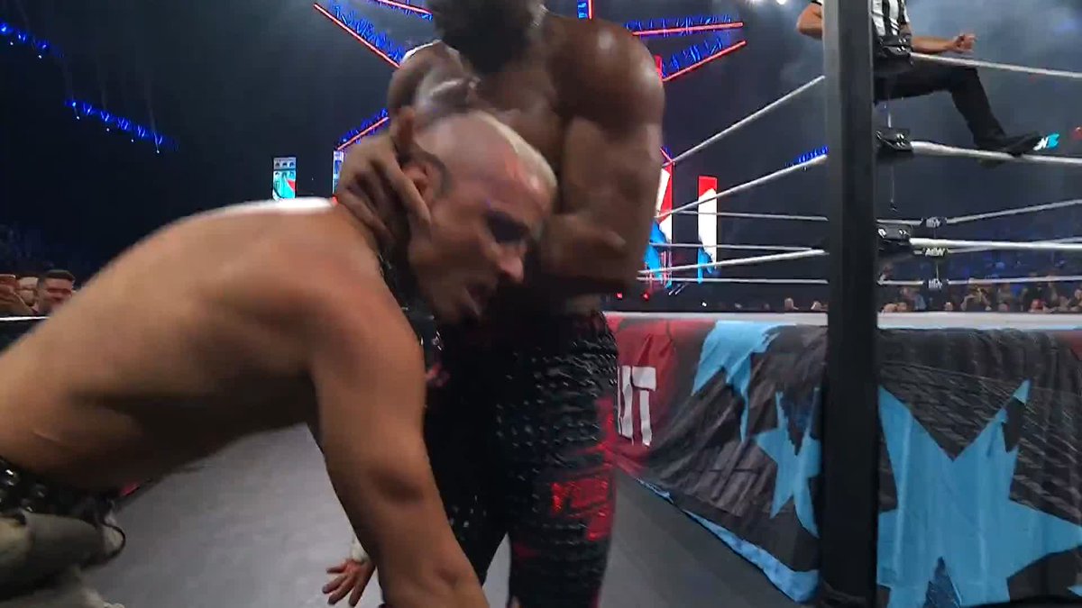 A wrestler in black and red gear grapples a bald opponent against the ring apron, with the AEW logo visible on the barricade, during the AEW All Out pay-per-view event broadcasting live on HBO Max PPV.