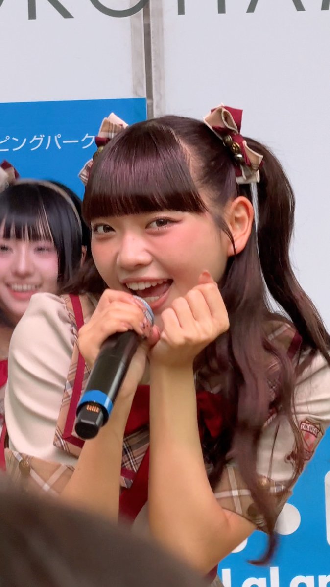 A smiling young woman with dark hair in pigtails and plaid bows speaks into a microphone at an event, with partial Japanese text visible in the background.