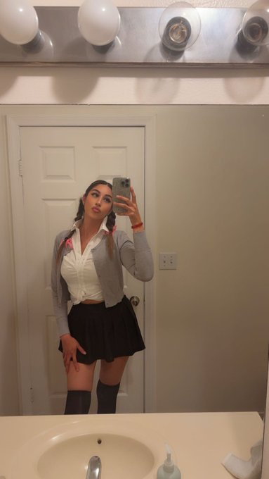 A young woman takes a mirror selfie in a bathroom wearing a private school-inspired outfit of a white button-up, gray cardigan, black pleated skirt, and over-the-knee socks, evoking the iconic "...Baby One More Time" aesthetic famously worn by Britney Spears.