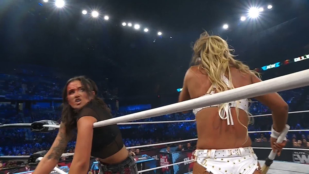 Two female wrestlers face off inside a brightly lit wrestling ring, with the woman on the right in a studded white outfit turning her back to the camera while her dark-haired opponent in black stares her down with an intense, defiant expression, capturing the heated confrontation referenced in the promotional quote. Partial signage visible in the background includes "AEW" and "ALL OUT" branding, tying the scene to the AEW All Out pay-per-view event.