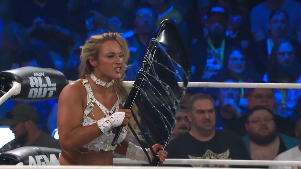 A blonde female wrestler in a white studded outfit grips a chair wrapped in barbed wire inside the ring at AEW All Out, as the crowd watches in the background. The "AEW All Out" branding is visible on the turnbuckle padding to the left, setting the stage for the dangerous maneuver described in the caption.