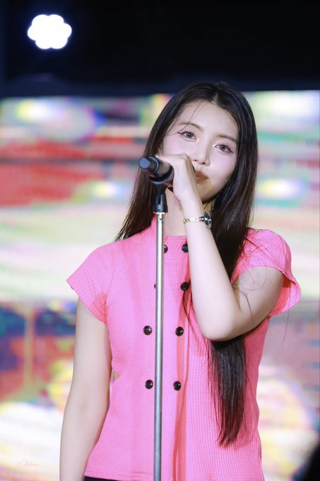 A young woman with long dark hair in a pink short-sleeved top stands at a microphone, appearing to perform on stage.