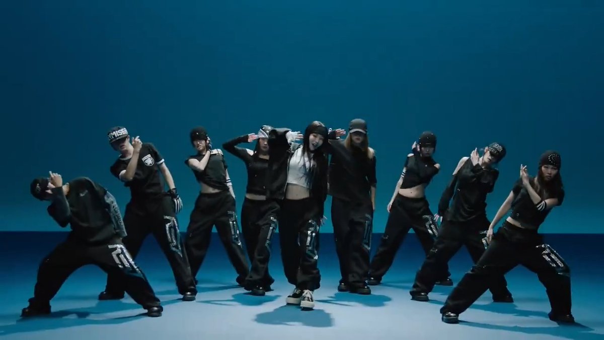 A group of female K-pop dancers in black streetwear outfits performing a synchronized choreography on a blue stage with teal background.